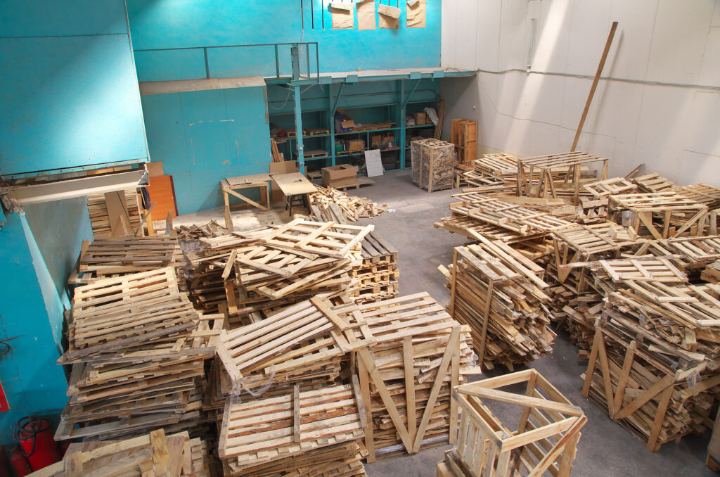 A dump of pallets in a warehouse with blueish/green and white walls.